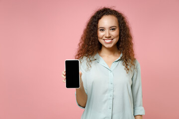 Wall Mural - Young black african american smiling curly woman 20s wearing casual blue shirt hold in hand using mobile cell phone with blank screen workspace area isolated on pastel pink background studio portrait.