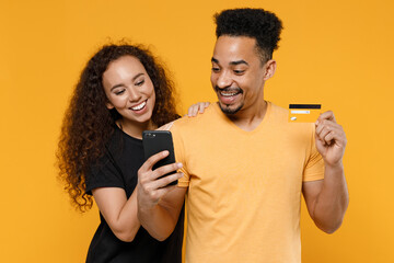 Wall Mural - Young couple two friends together family african smiling happy man woman 20s in black t-shirt hold mobile cell phone credit bank card shop online isolated on yellow color background studio portrait