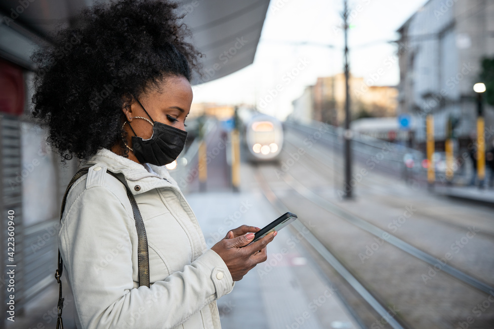Young woman in the evening at sunset in city wears protective face mask during the global pandemic from Covid-19 Coronavirus looks at the smartphone while waiting for the tram to go home after work - obrazy, fototapety, plakaty 