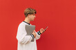 Handsome young man in stylish street clothes stands on a background of a red wall in wireless headphones, laptop and smartphone in his hands, looking away with a serious face on copy space