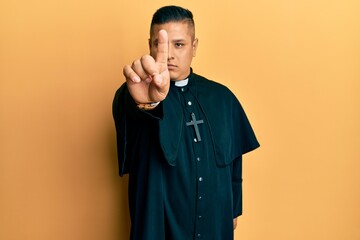 Young latin priest man standing over yellow background pointing with finger up and angry expression, showing no gesture