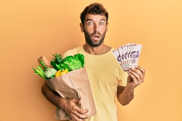 Poster - Handsome man with beard holding groceries and 500 mexican pesos banknotes in shock face, looking skeptical and sarcastic, surprised with open mouth