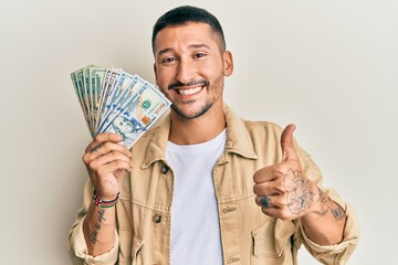 Poster - Handsome man with tattoos holding dollars smiling happy and positive, thumb up doing excellent and approval sign