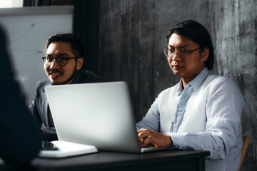 Canvas Print - Two Vietnamese men in business suits and glasses are sitting in the office at a table with a laptop and conducting negotiations. Brainstorming concept, office work