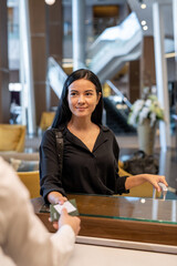 Poster - Happy young businesswoman or female traveler with luggage standing by reception counter and passing her documents to receptionist