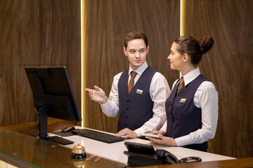 Aufkleber - One of two young hotel receptionists standing by counter, looking at colleague with touchpad and pointing at computer screen