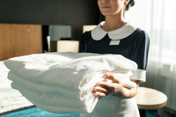 Poster - Hand of young beautiful chambermaid in uniform carrying stack of clean folded linens or towels while standing against in hotel room