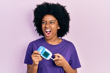 Poster - Young african american woman holding glucometer device angry and mad screaming frustrated and furious, shouting with anger. rage and aggressive concept.