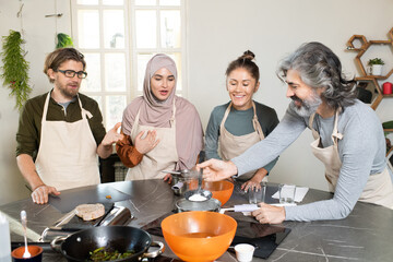 Wall Mural - Happy mature bearded man holding kitchen tool over metallic pan with boiling water while cooking by table among learners of masterclass