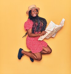 Canvas Print - Young beautiful african american woman wearing summer clothes and hat smiling happy. Jumping with smile on face holding city map over isolated yellow background.