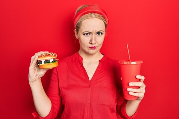 Canvas Print - Young blonde woman eating a tasty classic burger with fries and soda skeptic and nervous, frowning upset because of problem. negative person.