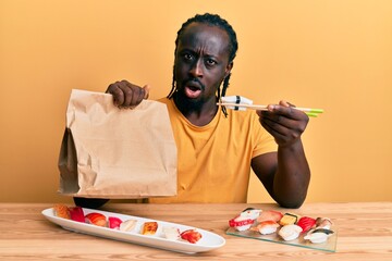 Sticker - Handsome young black man eating take away sushi sitting on the table in shock face, looking skeptical and sarcastic, surprised with open mouth