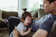 © Maskot - Father on paternity leave looking at male toddler crying in living room