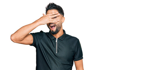 Canvas Print - Young man with beard wearing sportswear peeking in shock covering face and eyes with hand, looking through fingers with embarrassed expression.