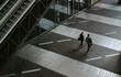 © Maskot - Rear view of male and female colleagues walking by escalator on walkway