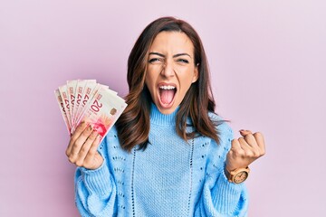 Poster - Young brunette woman holding 20 israel shekels banknotes angry and mad raising fist frustrated and furious while shouting with anger. rage and aggressive concept.