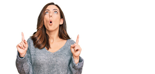 Young brunette woman wearing casual winter sweater amazed and surprised looking up and pointing with fingers and raised arms.