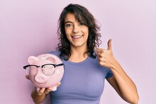 Young Hispanic Woman Holding Piggy Bank With Glasses Smiling Happy And Positive, Thumb Up Doing Excellent And Approval Sign