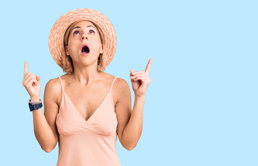 Canvas Print - Young blonde woman wearing summer hat amazed and surprised looking up and pointing with fingers and raised arms.