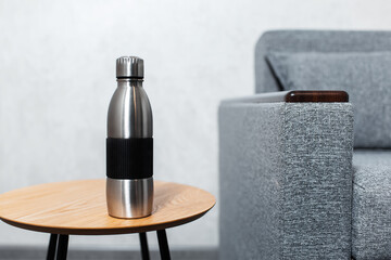 Close-up of steel reusable thermo water bottle on wooden table against grey wall near sofa.