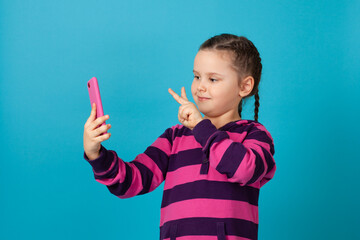 close-up portrait of a smiling girl with pigtails talking on a phone video chat and showing a victory sign with two fingers, isolated on a blue background.