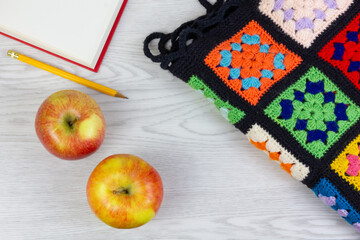 Above view of two yellow and red apples, a multicolor plaid blanket, an open book and a pencil on a white wooden table