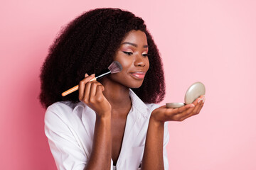 Sticker - Photo of charming dark skin lady applying shadow look hand hold little mirror isolated on pink color background