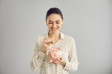 Happy beautiful independent young woman putting coin in piggy bank and smiling standing in studio with light grey background. Earning money, saving up, managing finances and planning budget concept