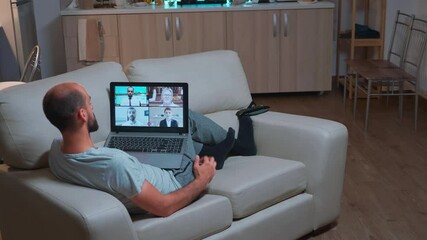 Poster - Caucasian male talking about business communication project during video call meeting use laptop computer. Focused man lying on sofa in pijamas using modern technology wireless late at night