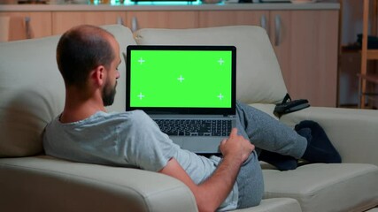 Poster - Tired man lying on sofa working on computer with mock up green screen chroma key display searching online information. Caucasian male using isolated pc late at night in kitchen