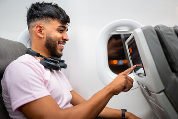 Passenger on the plane touching the LCD entertainment screen. Latin american man in airplane cabin, using smart device, with headphones.