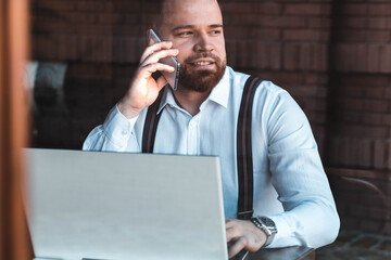 Young hipster, businessman talking over a phone in a cafe, laptop on the table