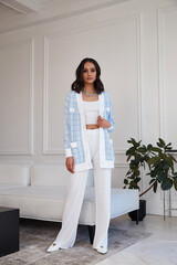 Wall Mural - Pretty fashion beautiful woman sexy lady brunette curly hair dark tanned skin wear trend clothes knitted suit blue jacket white top pants shoes interior room sofa plants spring collection.