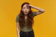 Surprised, positive woman with long ginger hair. Wearing green t-shirt. People and emotion concept. Gaze into distance with palm over her eyes. Watching at the camera, isolated over orange background