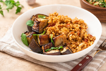 Wall Mural - Bulgur pilaf (pilavi) with tomatoes and fried eggplants in a white plate on a light table close-up. Turkish cuisine, vegan food.