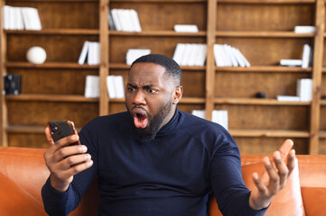 Shocked African-American man holding a smartphone, feel astonishment with a bad news, fired from work, irritated guy looks at phone screen and does not understand what happened