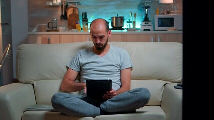 Poster - Tired man sitting alone on couch while browsing on internet using tablet computer with modern technology wireless. Caucasian male in pajamas relaxing in front of televion late at night in kitchen