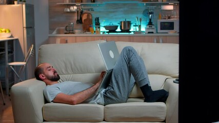 Poster - Exhausted man falling asleep while working on internet communication project using laptop computer with modern technology wireless. Caucasian male in pajamas sitting on sofa late at night in kitchen