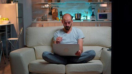 Poster - Tired man in pajams browsing on internet lifestyle information using laptop computer sitting on sofa. Freelancer working at online project in front on television late at night in kitchen