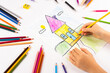 Child girl draws house with multicolored felt-tip pens on a white sheet.