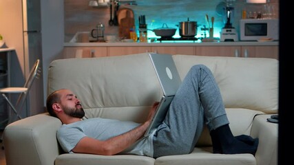 Poster - Man falling asleep in fron of TV while working on the laptop. Cozy man lying down on sofa while typing on laptop computer for social media project. Exhausted man working