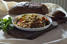 Pasta With Vegetables On A White Plate Stands On A Wooden Textured Plank And Textiles, In The Background Is Soddy Rustic Bread And Cilantro
