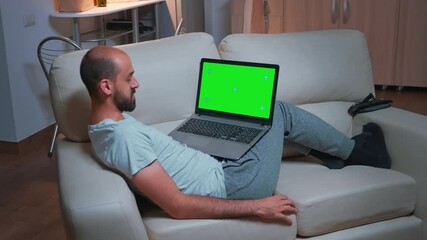 Poster - Tired man sitting on sofa while browsing marketing information using laptop computer with mock up green screen chroma key display. Caucasian male using modern technology wireless late at night