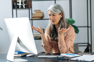 Outraged annoyed beautiful Asian mature woman, business lady or manager, looks at computer in confusion, shouts, receives unexpected news, fails the deal, gestures, sits at the table in the office