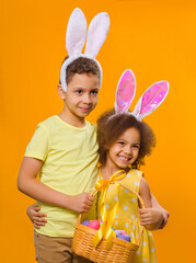 Wall Mural - A cheerful black girl and boy with rabbit ears on her head with a basket of colored eggs in her hands at Easter holyday