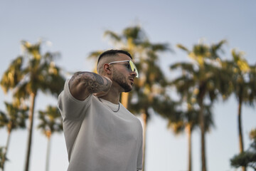 Wall Mural - Portrait of a tattooed young man wearing stylish sunglasses and posing against palm trees