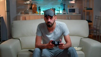 Wall Mural - Front view of pro gamer sitting on sofa in front of television while playing videogames competition using wireless joystick. Cheerful man with sleep mask enjoying time alone late at night in kitchen