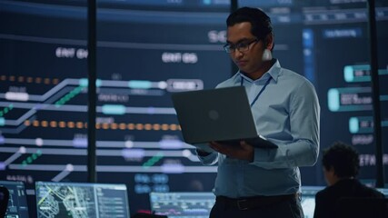 Aufkleber - Young Multiethnic Male Government Employee Uses Laptop Computer in System Control Monitoring Center. In the Background His Coworkers at Their Workspaces with Many Displays Showing Technical Data.