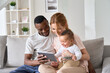 © insta_photos - Happy young multiracial family couple holding cute infant daughter having fun with digital tablet sitting on sofa. Funny mixed race child baby girl laughing using computer with diverse parents at home