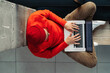 © Yury - Above view of young freelancer man in glasses wear red hat and wool sweater sitting on bench next by window legs crossed, remote online working on laptop, typing on keyboard. Distance job.